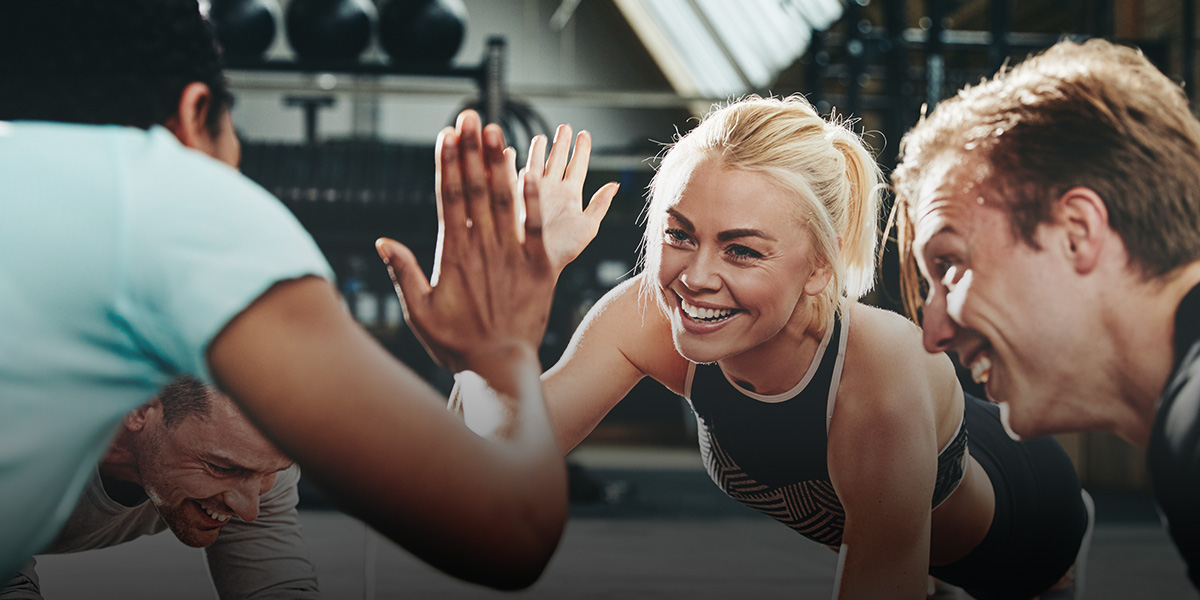 Three adults exercising together on a gym floor, smiling and giving a high-five while holding a plank position.